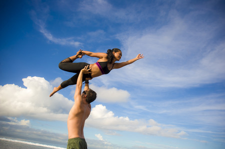 young attractive and concentrated couple of acrobats practicing acro yoga balance and meditation exercise on beautiful beach under a blue sky in mind and body control and healthy lifestyleの写真素材
