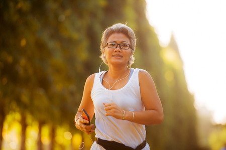 middle aged 40s or 50s happy and attractive woman with grey hair training at city park with green trees on sunrise doing running and jogging workout in health care fitness and healthy lifestyleの写真素材