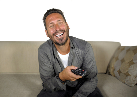young attractive and happy man laughing at home watching television show or comedy series holding remote sitting at living room sofa couch relaxed and cheerful in domestic entertainment lifestyleの写真素材