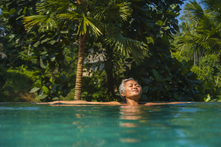 natural lifestyle portrait of attractive and happy middle aged Asian woman relaxed at tropical resort infinity swimming pool with jungle background enjoying relaxed in luxury holidays tripの写真素材