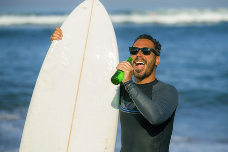 natural portrait of attractive and happy surfer man on his 40s carrying surf board after surfing morning at beautiful beach in wetsuit drinking beer bottle  enjoying Summer holiday and sport lifestyleの写真素材