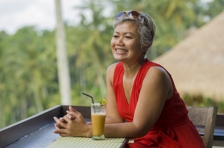 natural lifestyle outdoors portrait of attractive and happy middle aged Asian Indonesian woman in stylish red dress enjoying tropical holiday trip at tourist resort with beautiful jungle viewの写真素材