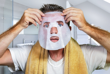lifestyle bathroom portrait of young scared and surprised man at home using beauty paper facial mask cleansing doing anti aging facial treatment looking himself weird and funny in toilet mirrorの写真素材