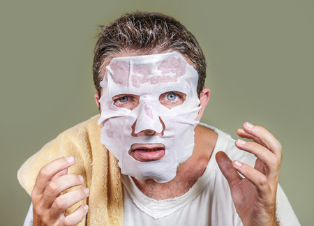 lifestyle home portrait of young funny and messy man applying beauty facial mask looking to the mirror surprised and horrified in skin care aging concept and male cosmetic face treatmentの写真素材