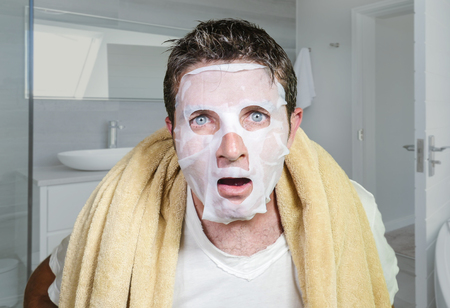 lifestyle home portrait of young funny and messy man applying beauty facial mask looking to the mirror surprised and horrified in skin care aging concept and male cosmetic face treatmentの写真素材