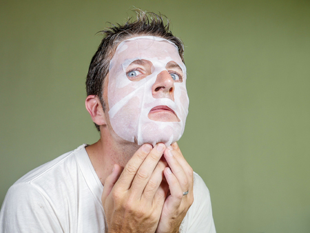 lifestyle isolated background portrait of young weird and funny man at home trying using beauty paper facial mask cleansing learning anti aging treatment in concentrated face expressionの写真素材