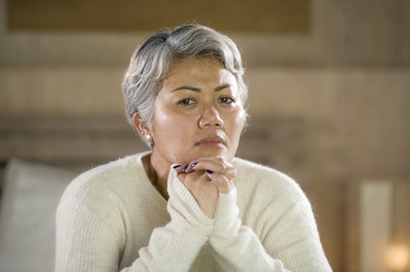 Depressed 40s - 50s mature female, dramatic lifestyle home portrait of attractive sad and lost middle aged woman with grey hair sitting on bed feeling frustrated suffering depression and painの写真素材