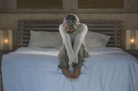 Depressed 40s - 50s mature female, dramatic lifestyle home portrait of attractive sad and lost middle aged woman with grey hair sitting on bed feeling frustrated suffering depression and painの写真素材