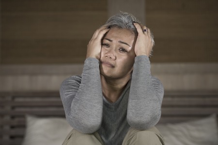 Depressed 40s - 50s mature female, dramatic lifestyle home portrait of attractive sad and lost middle aged woman with grey hair sitting on bed feeling frustrated suffering depression and painの写真素材