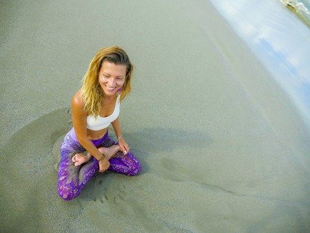 young happy and attractive blond girl doing yoga and concentration exercise outdoors at beautiful beach in relax and meditation practice sitting in lotus position in wellness and healthy lifestyleの写真素材