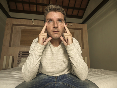 dramatic home portrait of 30s to 40s attractive depressed and desperate man sitting on bed suffering anxiety crisis and depression problem crying sad and thoughtful feeling overwhelmed and lostの写真素材