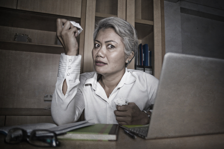 Attractive desperate and stressed middle aged Asian woman screaming gesturing overwhelmed and overwork working at office computer desk feeling exploited and upset in work stressの写真素材
