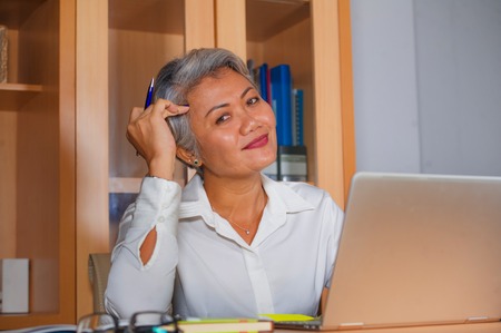 lifestyle office portrait of attractive and happy successful middle aged Asian woman working at laptop computer desk smiling confident in entrepreneur business and financial successの写真素材