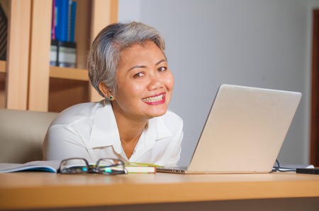 natural lifestyle office portrait of attractive and happy successful mature Asian woman working at laptop computer desk smiling confident in entrepreneur business financial successの写真素材