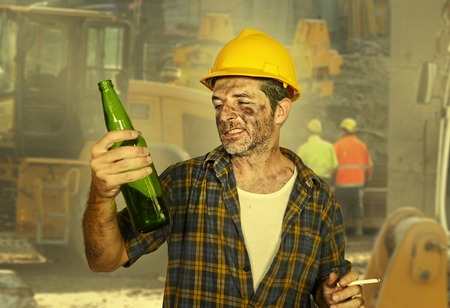 lifestyle portrait of thirsty and tired constructor worker or builder man in safety helmet feeling exhausted drinking cold beer bottle refreshing during work break at building site smiling happyの写真素材