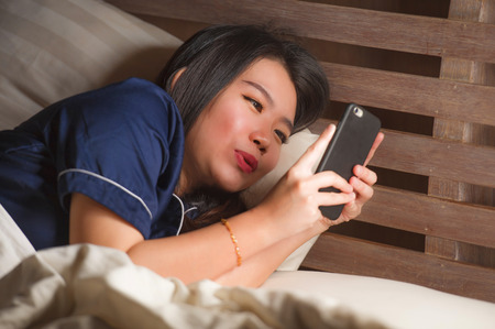 young beautiful and happy Asian Korean woman in pajamas using mobile phone social media texting with her boyfriend or enjoying online dating app smiling cheerful lying on bed at homeの写真素材