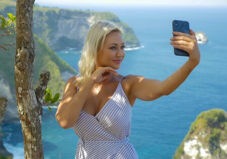 young happy and attractive blond woman taking selfie portrait with mobile phone at beautiful tropical paradise view of ocean rock cliff enjoying Summer holidays in travel and tourist conceptの写真素材