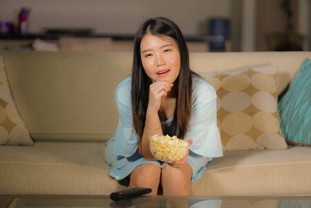 young happy and excited Asian Chinese woman with TV remote controller eating popcorn bowl watching television enjoying Korean drama or comedy movie having fun relaxed and cheerfulの写真素材