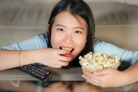 young happy and excited Asian Chinese woman with TV remote controller eating popcorn bowl watching television enjoying Korean drama or comedy movie having fun relaxed and cheerfulの写真素材