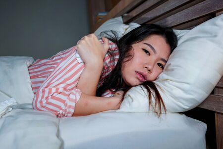 young beautiful sad and depressed Asian Korean woman trembling in bed suffering cold and flu at night feeling unwell at night in her bedroom in health care and depression problem conceptの写真素材