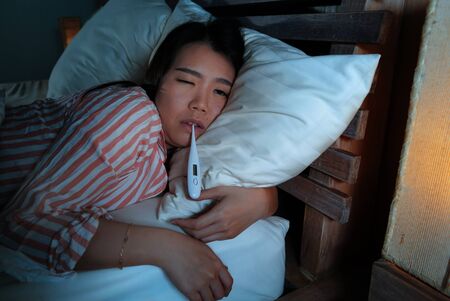 young beautiful sick and exhausted Asian Chinese woman suffering cold and flu taking temperature with thermometer lying on bed at night feeling ill and unwell in health care and gripe conceptの写真素材