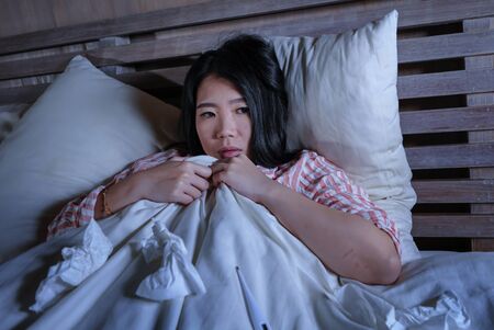 young beautiful tired and sick Asian Japanese woman lying ill on bed in pajamas at night with tissue papers suffering cold and flu feeling unwell and exhausted in health care and winter virus conceptの写真素材