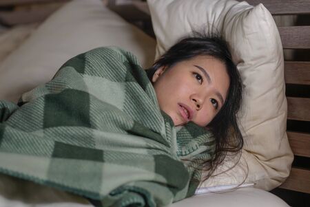 lifestyle portrait of young beautiful sick and exhausted Asian Chinese woman suffering cold and flu having temperature lying on bed covering with blanket feeling ill and unwell awake at nightの写真素材