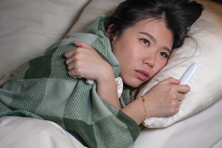 young beautiful tired and ill Asian Korean woman lying on bed at home sick suffering cold flu and temperature covered with blanket feeling unwell and feverish in medical care and health conceptの写真素材