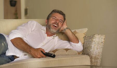 home lifestyle portrait of young attractive happy and cheerful man watching television funny show or comedy movie laughing relaxed holding TV remote lying at living room sofa couchの写真素材
