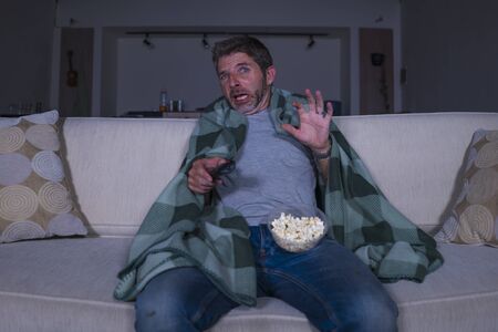 funny home lifestyle portrait of scared and frightened man alone at night in living room couch watching horror scary movie in television screaming and eating popcorn covering with blanketの写真素材