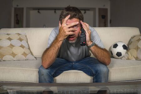 home lifestyle portrait of young attractive nervous and excited football supporter man watching soccer game on television at living room sofa couch feeling crazy intense emotion and stressの写真素材