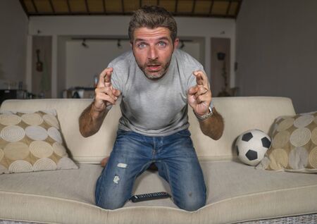 lifestyle portrait of young attractive nervous and excited football supporter man watching soccer game on television at living room couch feeling intense emotion and stress crossing fingersの写真素材