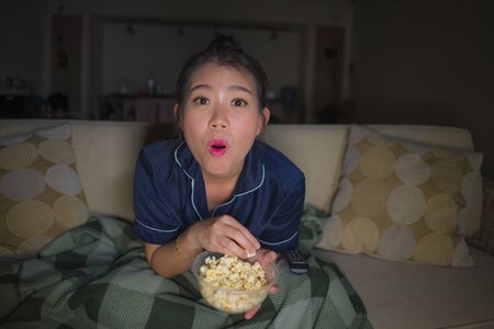 young beautiful happy and relaxed Asian Chinese woman at home living room sitting cozy on sofa couch watching TV show episode or romantic comedy movie eating popcorn smiling and having funの写真素材