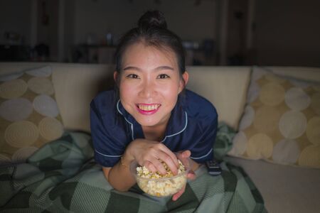 young beautiful happy and relaxed Asian Korean woman at home living room sitting cozy on sofa couch watching TV show episode or romantic comedy movie eating popcorn smiling and having funの写真素材
