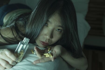 young beautiful desperate and wasted addict Asian Japanese woman taking drug overdose lying on bed feeling sick and depressed suffering depression breakdown passed out in pills addictionの写真素材