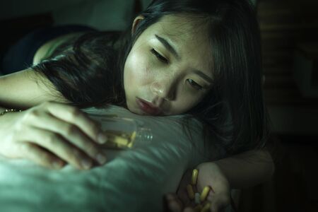 young beautiful desperate and wasted addict Asian Korean woman taking drug overdose lying on bed feeling sick and depressed suffering depression breakdown passed out in pills addictionの写真素材