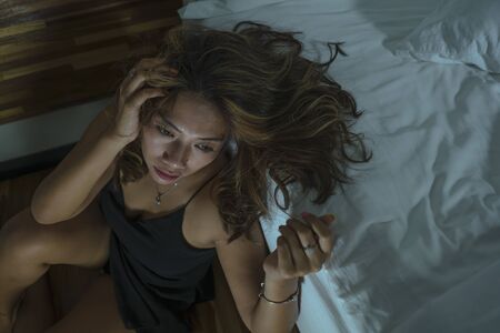 dramatic home lifestyle portrait of young beautiful sad and depressed Asian Indonesian woman in nightgown on bedroom floor by the bed feeling broken and lost suffering depression and anxietyの写真素材