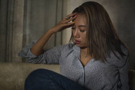 dramatic portrait of young beautiful sad and depressed Asian woman in pain thoughtful and confused at home couch feeling broken heart suffering depression crisis and anxiety problemの写真素材