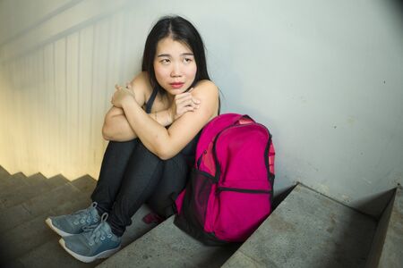 Dramatic portrait of Asian female college student bullied. Young depressed and sad Chinese girl sitting lonely on campus staircase suffering bullying and harassment feeling desperate and excludedの写真素材