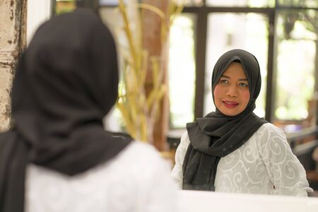 lifestyle portrait of elegant and happy middle aged Asian Indonesian Muslim woman in traditional hijab adjusting the Islamic scarf in front of the mirrorの写真素材