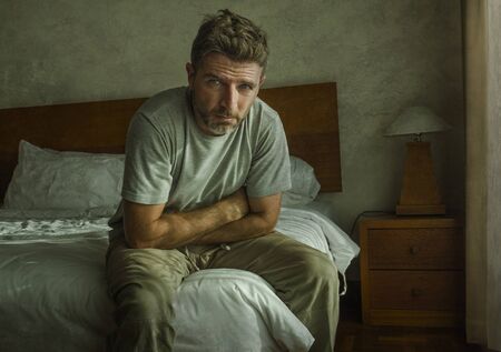 Attractive depressed and upset man at home bedroom . dramatic lifestyle portrait of 30s to 40s handsome guy sitting sad on bed feeling worried and desperate suffering depression problemの写真素材