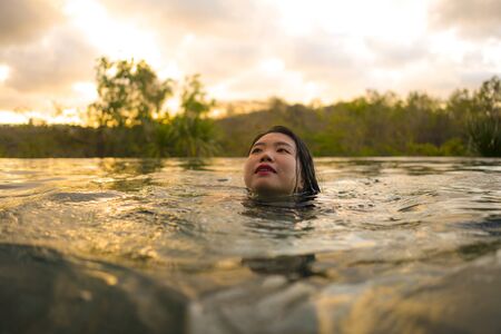 tropical holidays lifestyle portrait of young beautiful and happy Asian Chinese woman in bikini enjoying sunset at amazing jungle resort infinity pool swimming relaxed and carefree in Summer vacationの写真素材