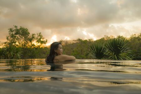 tropical holidays lifestyle portrait of young beautiful and happy Asian Chinese woman in bikini enjoying sunset at amazing jungle resort infinity pool swimming relaxed and carefree in Summer vacationの写真素材
