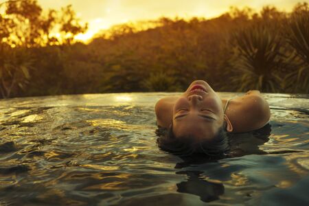 tropical holidays lifestyle portrait of young beautiful and happy Asian Chinese woman in bikini enjoying sunset at amazing jungle resort infinity pool swimming relaxed and carefree in Summer vacationの写真素材