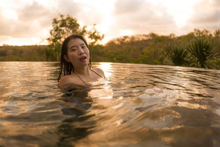 tropical holidays lifestyle portrait of young beautiful and happy Asian Chinese woman in bikini enjoying sunset at amazing jungle resort infinity pool swimming relaxed and carefree in Summer vacationの写真素材