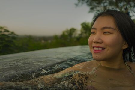tropical holidays lifestyle portrait of young beautiful and happy Asian Korean woman in bikini enjoying sunset at amazing jungle resort infinity pool swimming relaxed and carefree in Summer vacationの写真素材