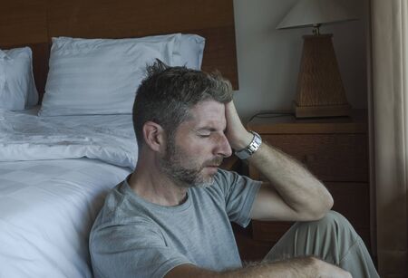 dramatic home portrait of young depressed and desperate man sitting on bedroom floor next to bed suffering depression and anxiety feeling overwhelmed and helplessの写真素材