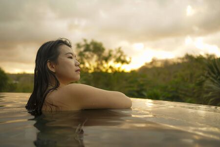 tropical holidays lifestyle portrait of young beautiful and happy Asian Korean woman in bikini enjoying sunset at amazing jungle resort infinity pool swimming relaxed and carefree in Summer vacationの写真素材