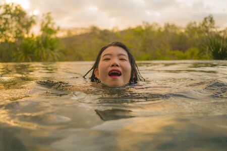 tropical holidays lifestyle portrait of young beautiful and happy Asian Korean woman in bikini enjoying sunset at amazing jungle resort infinity pool swimming relaxed and carefree in Summer vacationの写真素材