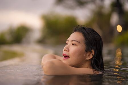 tropical holidays lifestyle portrait of young beautiful and happy Asian Korean woman in bikini enjoying sunset at amazing jungle resort infinity pool swimming relaxed and carefree in Summer vacationの写真素材